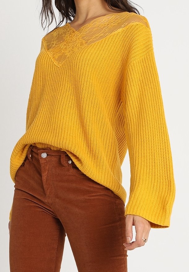 Żółty sweter dziany z koronkowym dekoltem, zestawiony z brązowymi sztruksowymi spodniami. Sweter ma luźny krój i długie rękawy.