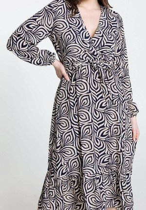 Kobieta w długiej sukience z długim rękawem i dekoltem w serek, w czarno-beżowy abstrakcyjny wzór liści, z paskiem podkreślającym talię i falbaną na dole.