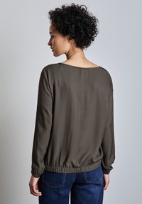 Olivgrünes Blusenshirt mit langen Ärmeln, elastischem Saum und weitem Ausschnitt hinten, getragen mit blauen Jeans. Glatte Textur und lockere Passform.