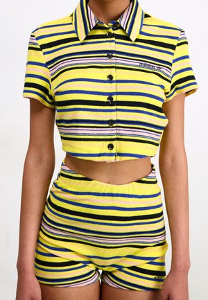 Frau trägt ein kurzärmeliges, knöpfbares Crop-Top mit gelben, schwarzen, blauen und pinken Streifen sowie passende High-Waist-Shorts mit Adidas-Logos.