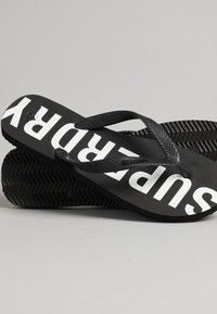 Tongs noires avec une semelle en caoutchouc texturé et de larges sangles arborant la marque "SUPERDRY" en blanc. Design classique et minimaliste avec une construction robuste.