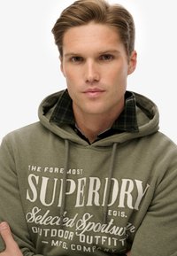 Sudadera verde oliva con un gráfico frontal y cordones, llevada sobre una camisa de cuadros en negro y verde. Textura suave y diseño casual.