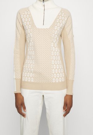 Beiger und weiß gemusterter Strickpullover mit gerippten Ärmeln und einem weißen Rollkragenpullover mit Reißverschluss, getragen mit weißen Hosen.