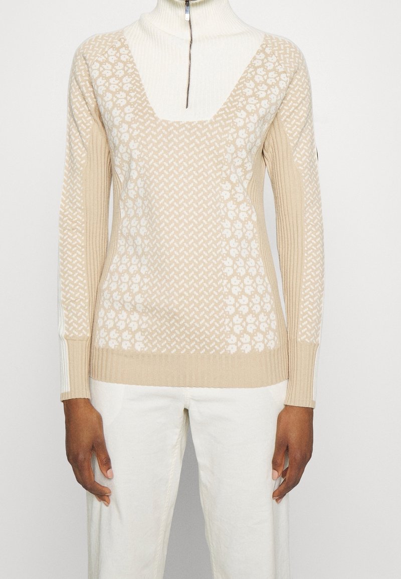 Pull en maille beige et blanc à motifs avec manches côtelées et col roulé blanc zippé, porté avec un pantalon blanc.