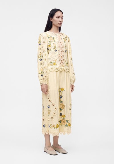 Woman standing wearing a cream-colored, floral embroidered skirt and long-sleeve top with lace details and beige flat shoes.