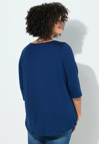 Navyblauw topje met driekwart mouwen, een ronde halslijn, zachte textuur en licht gebogen zoom. Draagt blauwe spijkerbroek en toont een tatoeage op de arm.