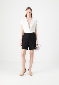 Hvit dyp v-hals bluse med cap sleeves, svarte skreddersydde shorts og sandaler med klare hæler. Holder en liten, lys rosa håndveske.