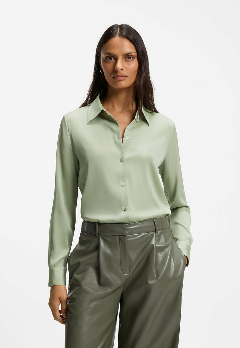 Femme aux longs cheveux foncés portant une chemise verte claire boutonnée et un pantalon en cuir vert foncé taille haute, les mains dans les poches.