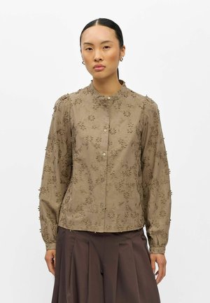 Kvinde med mørkt hår iført beige langærmet bluse med 3D blomsterbroderi og mørke plisserede bukser, stående mod en hvid baggrund.
