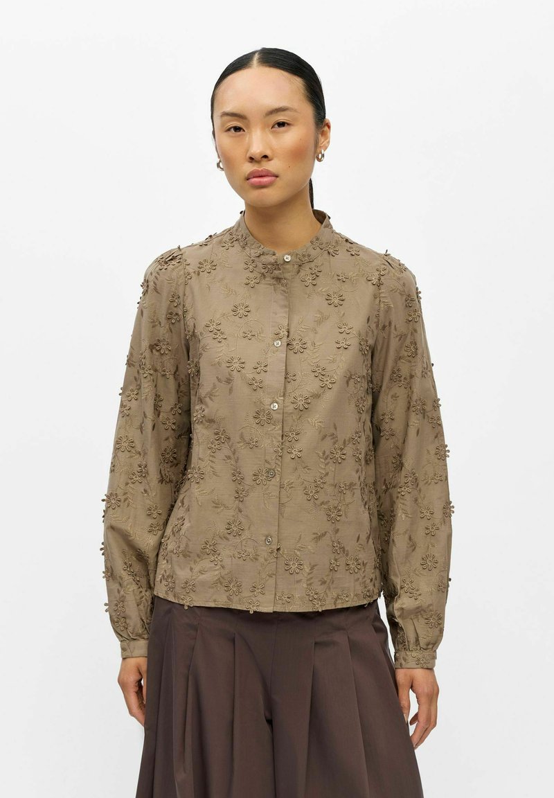 Femme aux cheveux foncés portant une blouse beige à manches longues avec broderie florale 3D et un pantalon plissé foncé, debout devant un fond blanc.