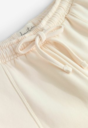 Shorts di cotone beige chiaro con vita elastica, dotati di un cordoncino e di un'etichetta con testo in corsivo. Texture liscia e design semplice.