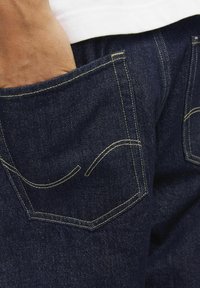 Mörkblå denimjeans med gul sömnad, som har en distinkt bakficka-design och en hand som vilar inuti fickan.