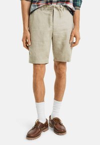 Beige Leinen-Shorts mit einem Kordelzugbund. Getragen mit weißen Socken und braunen Lederschuhen mit goldenen Hardware-Akzenten.