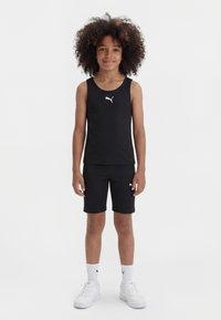 Παιδί με σγουρά μαλλιά που φοράει μαύρο τοπ και σορτς Puma, λευκές κάλτσες Puma και λευκά αθλητικά παπούτσια, στέκεται μπροστά σε λευκό φόντο.