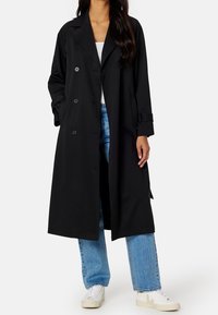 Černý trenčkot s dvouřadým zapínáním, dlouhými rukávy, bočními kapsami a volným střihem, kombinovaný se světlými modrými džínami a bílými botami.