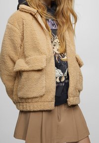 Veste en fausse fourrure beige avec fermeture éclair, deux poches avant et coupe ample, portée par-dessus un sweat à capuche graphique sombre et une jupe beige.