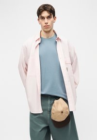 Νεαρός άνδρας με ανοιχτό ροζ πουκάμισο πάνω από μπλε μπλουζάκι και πράσινο παντελόνι, κρατώντας μπεζ και καφέ καπέλο κουμπωμένο στη μέση του.