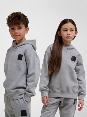 To børn iført matchende grå hættetrøjer og joggingbukser, stående side om side mod en ensfarvet baggrund.