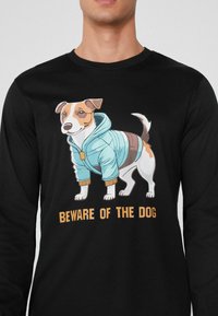Schwarz Langarmshirt mit einem Cartoon-Hund in einem hellblauen Kapuzenpulli mit orangefarbenen Akzenten. Darunter steht der Text: "VORSICHT VOR DEM HUND."