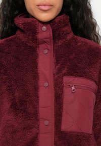 Burgundy fleece-takk med høy krage, trykknapper og en quiltet glidelåslomme. Strukturert stoff med myk, lodden utseende.