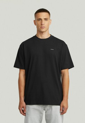 Uomo con capelli corti castani, che indossa una t-shirt nera oversize e pantaloni grigio chiaro, in piedi contro uno sfondo grigio chiaro.