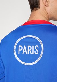 Plava sportska majica s crvenim ovratnikom na kojem se velikim, bijelim, zaobljenim tekstom nalazi riječ "PARIZ" unutar točkaste kružnice.