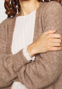 Brun stickad cardigan med en mjuk textur, med långa ärmar och en lös passform. Bärs över en vit topp med subtilt mönster och volangdetaljer vid ärmarna.