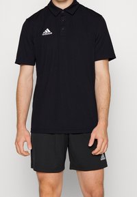 Czarny polo z miękkiej tkaniny z plisą na trzy guziki i krótkimi rękawami. Zawiera białe logo Adidas na lewej piersi.
