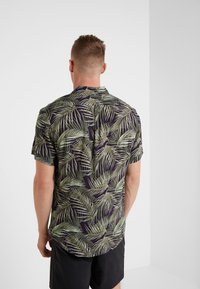 Mies joka on pukeutunut mustiin shortseihin ja lyhythihaiseen napitettavaan paitaan, jossa on vihreä trooppinen lehtikuosi, seisoo pelkää valkoista seinää vasten.