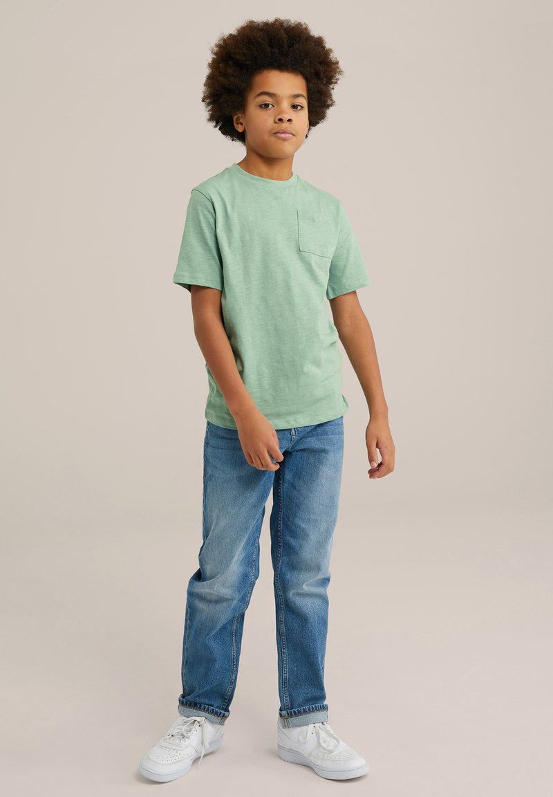 Svetle zelené tričko s krátkým rukávem a kapsou na prsou, nošené s modrými džíny a bílými teniskami, stojící na neutrálním pozadí.