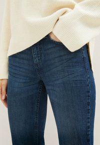 Mørkeblå denimjeans med smal passform, med subtil falming og klassisk femlommers design, parret med en cremegul ribbestrikket sweater.