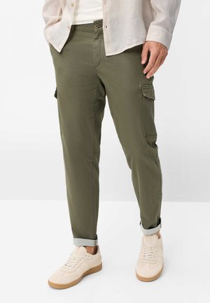 Mann trägt olivgrüne Cargo-Hosen mit aufgekrempelten Bündchen, beige Sneaker und ein hellbeiges Hemd über einem weißen Top.