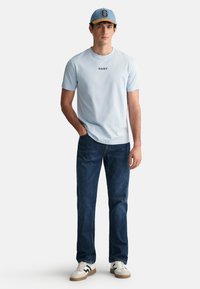 Tricou scurt, de culoare albastru deschis, cu logo-ul "GANT", asortat cu blugi de culoare albastru închis și adidași albi. Sapcă albastră cu o margine bej.