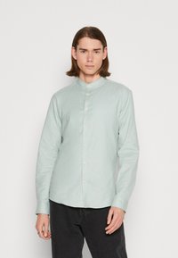 Camicia a maniche lunghe verde chiaro con colletto alla mandarino, chiusura con bottoni e un tessuto leggermente testurizzato, abbinata a pantaloni scuri.