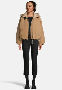 Beige bomberjack met een capuchon, ritssluiting en elastische manchetten. Draagt over een zwarte top en gecombineerd met zwarte broeken en veterschoenen.
