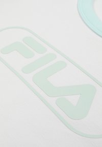 Gros plan d'un tissu blanc avec un logo FILA vert clair imprimé dessus, montrant une partie d'un motif circulaire en haut à droite.