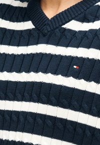 Pull rayé bleu marine et blanc avec un col en V, présentant un motif en maille torsadée texturée et un petit logo au centre.
