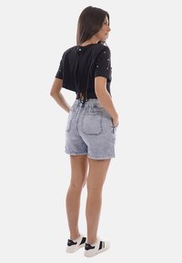 Donna con maglietta nera corta con dettagli in perle, pantaloncini di jeans chiari con tasche e scarpe da ginnastica bianche in piedi con le mani nelle tasche.