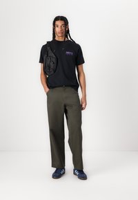 Schwarzes T-Shirt mit kurzen Ärmeln und lila Schriftzug, olivgrüne weite Hose und blaue Sneakers mit drei weißen Streifen. Umhängetasche über der Schulter getragen.