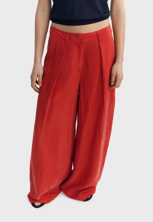 Pantalon rouge à jambes larges avec plis, bouton et passants pour ceinture, porté avec un haut noir et des chaussures noires à bout ouvert, sur fond uni.