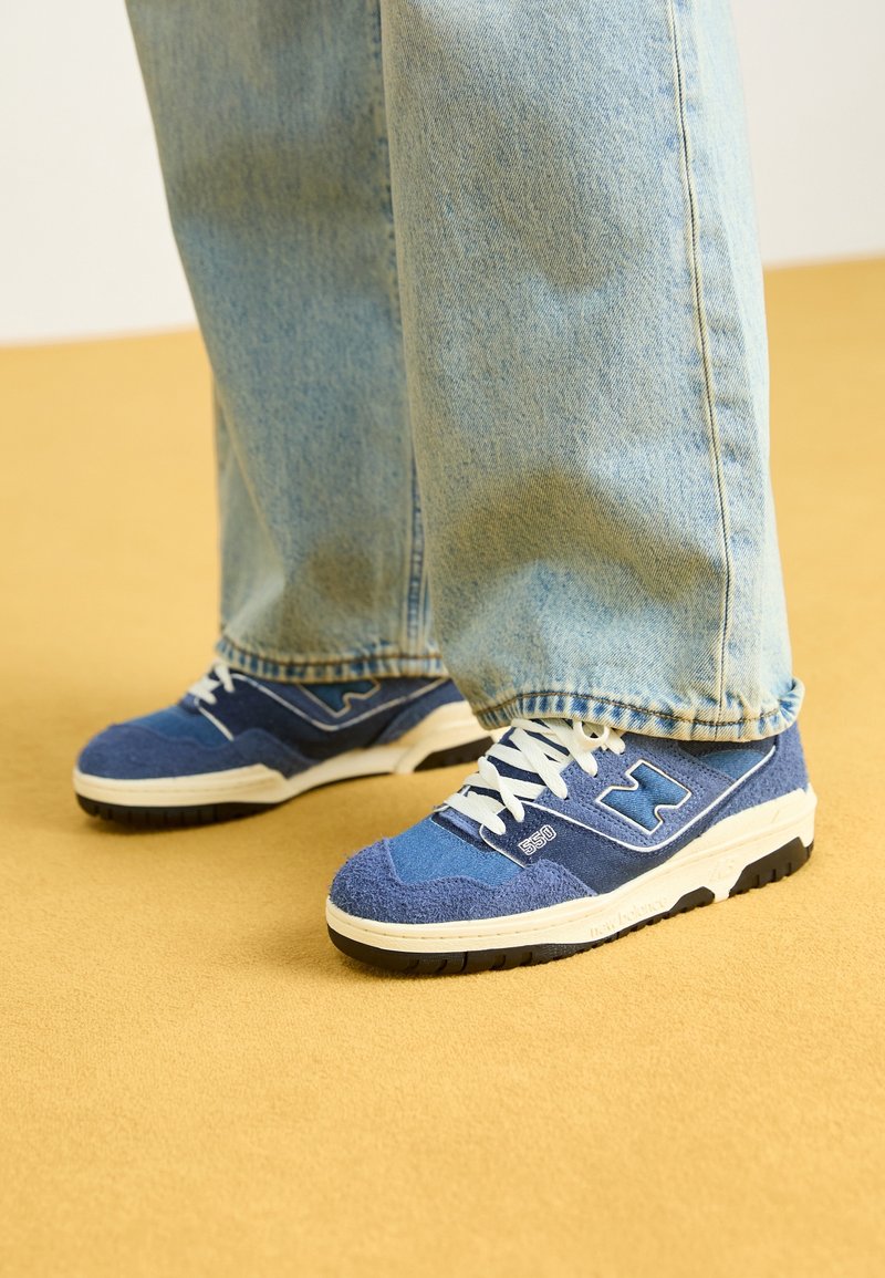 Jambes portant un jean bleu clair et des baskets en daim bleu avec des lacets blancs, debout sur un sol jaune.