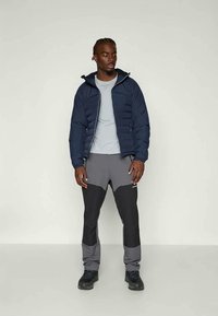 Veste rembourrée marine avec fermeture éclair et capuche, associée à un pantalon de sport à plusieurs panneaux gris. La tenue comprend un t-shirt gris ajusté et des chaussures noires.