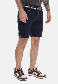 Bermuda blu navy con risvolti, una cintura a righe blu, marrone e bianca, e chiusura con bottone. Presenta una texture liscia e tasche laterali.