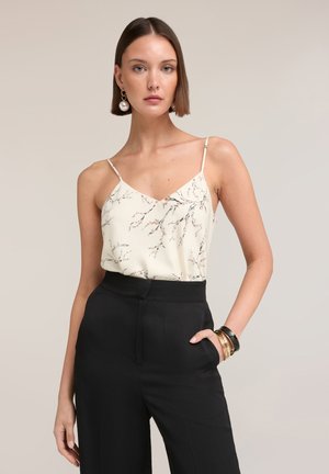 Donna con capelli corti castani che indossa una canotta bianca con fantasia floreale, pantaloni neri a vita alta, orecchino di perla e braccialetti, in piedi con una mano in tasca.