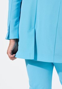 Giacca su misura azzurro chiaro con dettaglio sulla fessura, abbinata a pantaloni coordinati. Tessuto morbido con una leggera texture e polsini a quattro bottoni.