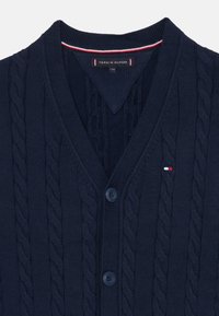 Cardigã tricotado azul marinho com padrão de tranças, decote em V e fecho de botão. Risca de destaque em vermelho e branco na gola. Textura suave.