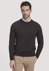 Donkergrijs gebreid sweater met een ronde halslijn, geribde manchetten en tailleband, gedragen over een lichtblauw overhemd met kraag, gecombineerd met beige broeken.