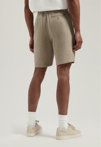 Beige, teksturerte shorts med elastisk linning og to baklommer. Modellen kombinerer dem med lyse joggesko og hvite sokker.