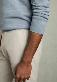 Pulover bleu deschis cu manșete texturate, asortat cu pantaloni bej deschis. Prim-plan pe braț și mână, evidențiind textura și contrastul de culori.