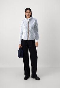 Giacca leggera azzurro chiaro con cappuccio, dotata di una chiusura centrale dorata e polsini elastici, abbinata a pantaloni neri a gamba larga.